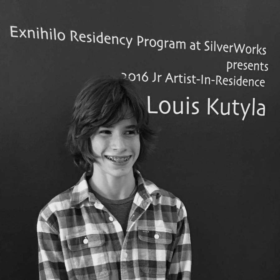 A smiling young person in a plaid shirt stands in front of a blue wall with event signage.