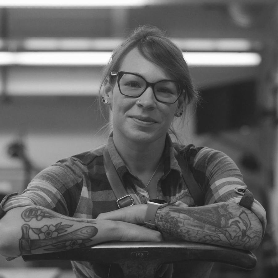 Photograph of a smiling woman with tattoos and glasses in a workshop.