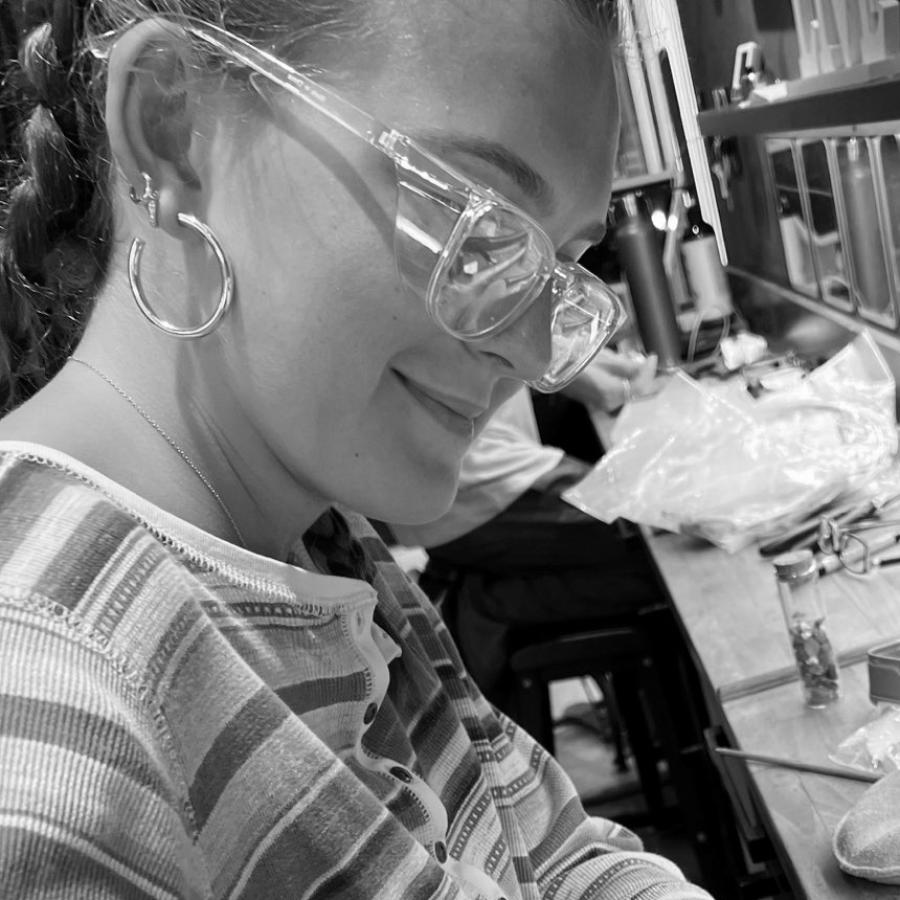 A young woman wearing safety glasses files a piece of metal at a workbench with jewelry tools.