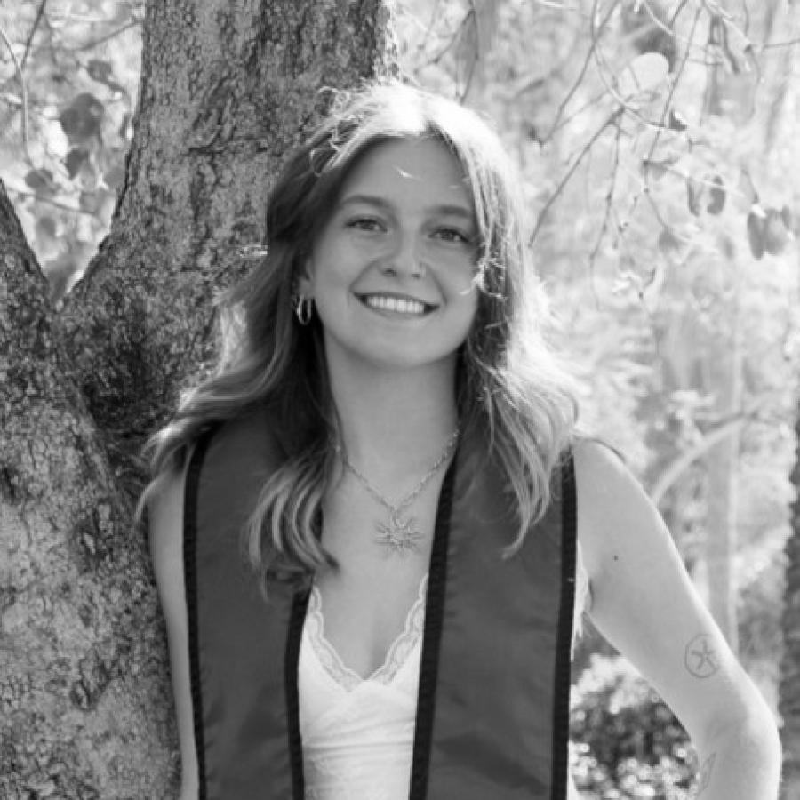 Photograph of a smiling graduate leaning against a tree wearing a red SDSU stole.