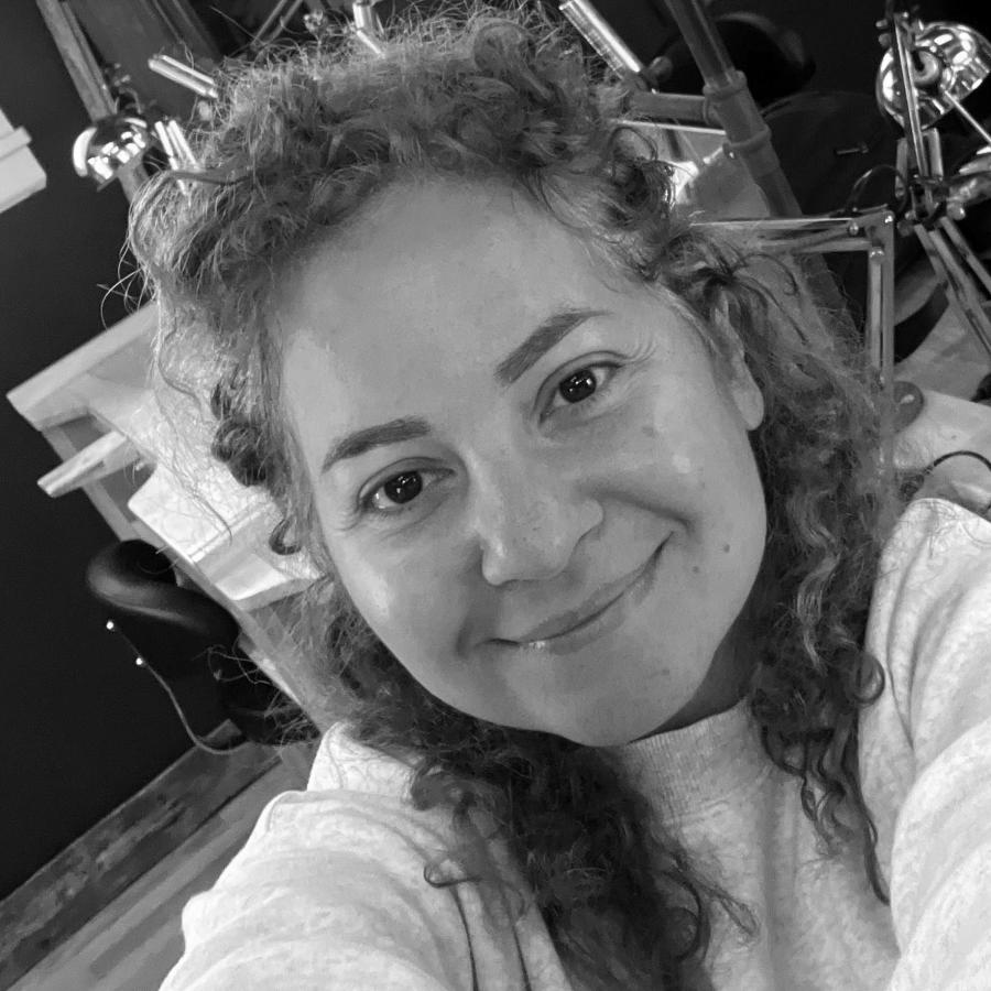 A smiling woman with curly hair takes a selfie in a jewelry workshop.