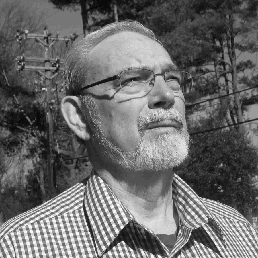 Photograph of a bearded older man wearing glasses and a blue checkered shirt outdoors.