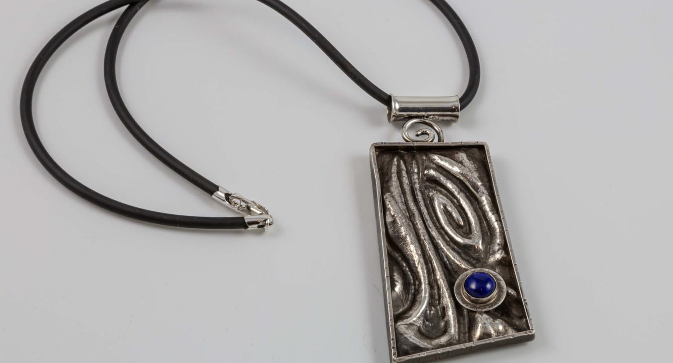 Photograph of a rectangular, textured silver pendant with a small blue stone on a black cord.
