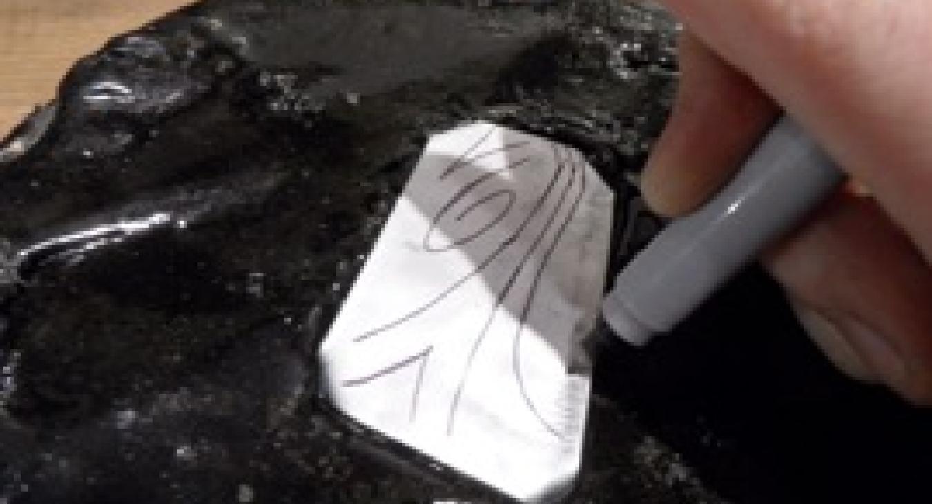 A close-up photograph of a person using a marker on a silver sheet on a black object.
