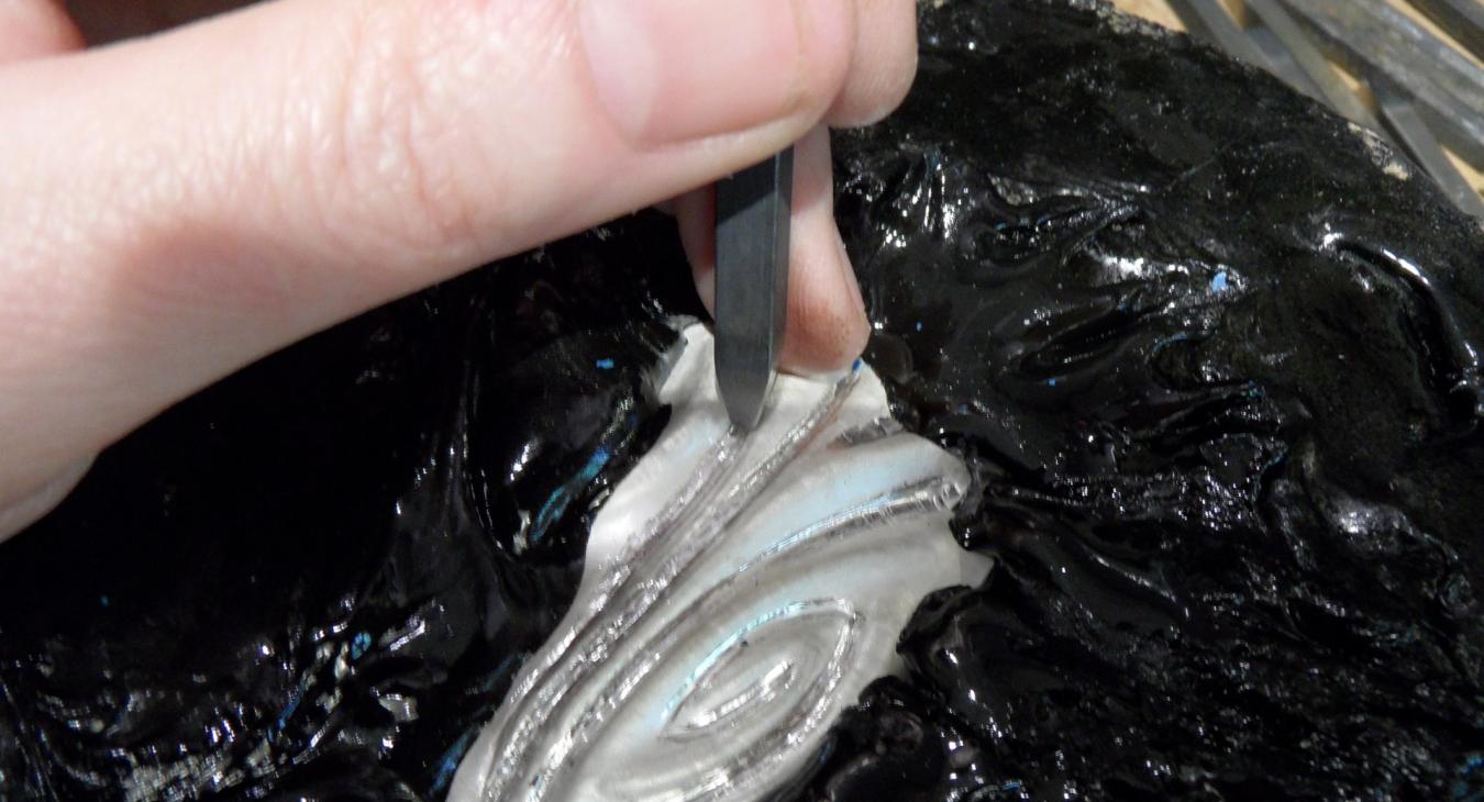 A close-up photograph of a hand using a graver to carve details into a piece of white mother-of-pearl secured in black pitch.