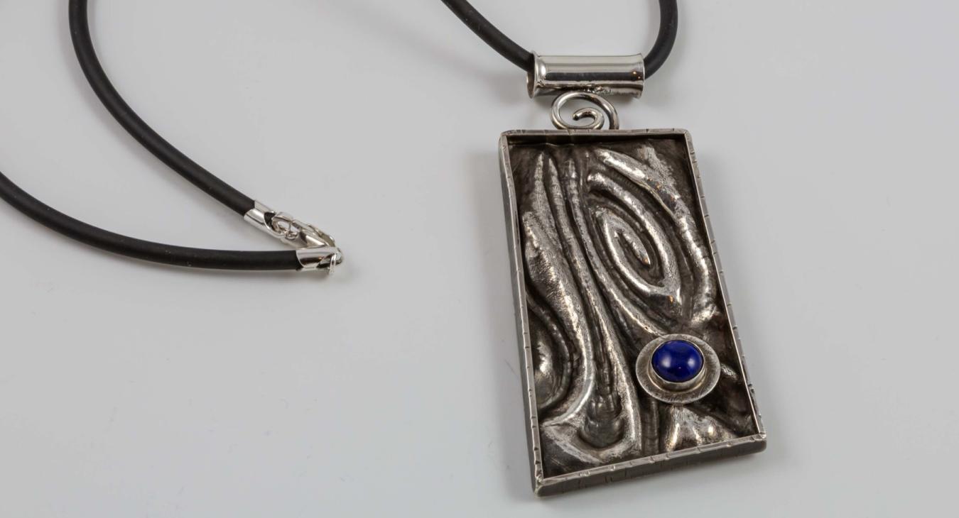 Photograph of a rectangular, textured silver pendant with a small blue stone on a black cord.