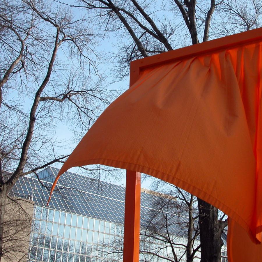 Orange fabric art blowing in wind with trees and building.