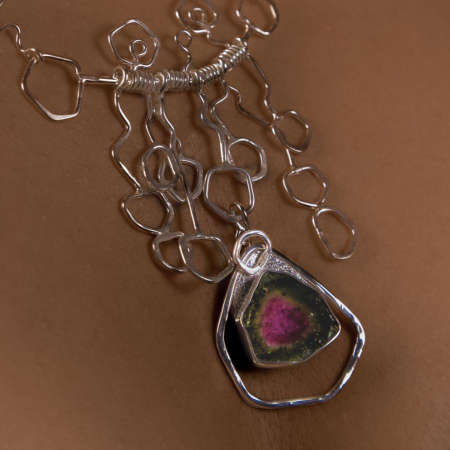 Model wearing a geometric necklace with repeated 5 sided shapes and a five sided pink and green stone