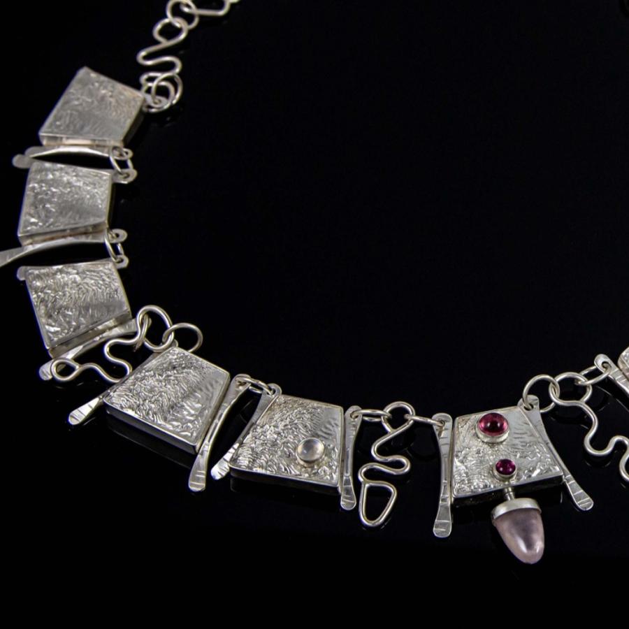 Photograph of a silver necklace with textured rectangular links, two red stones, and a pink stone.