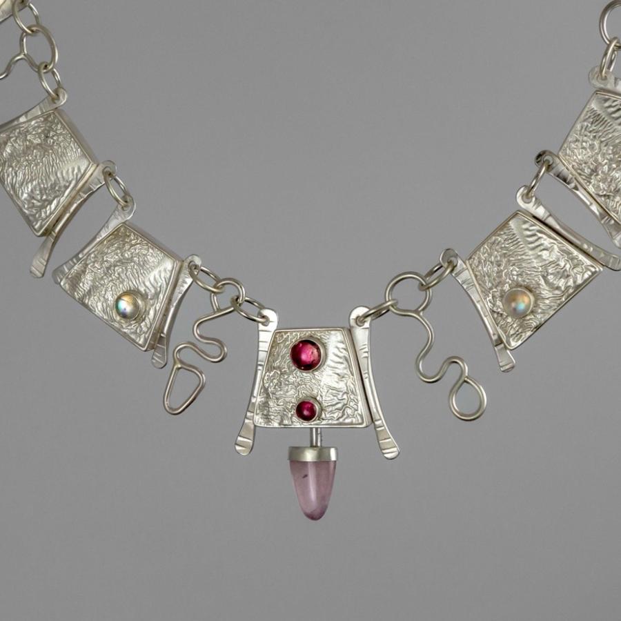 Photograph of a silver necklace with textured rectangular links, two red stones, and a pink stone.