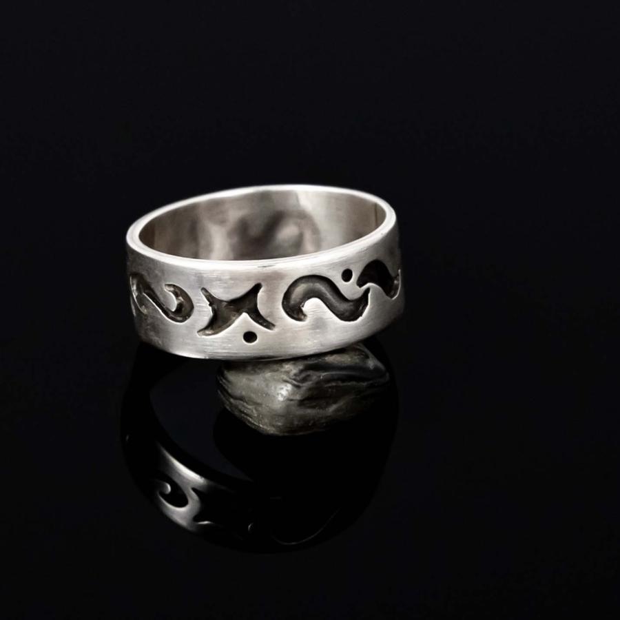 Photograph of a silver band ring with recessed symbols, reflected on a black surface.