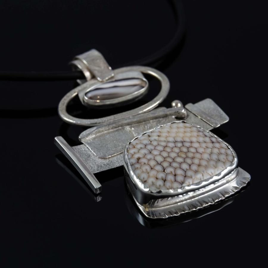 A silver pendant with a textured design on a black cord against a black background.