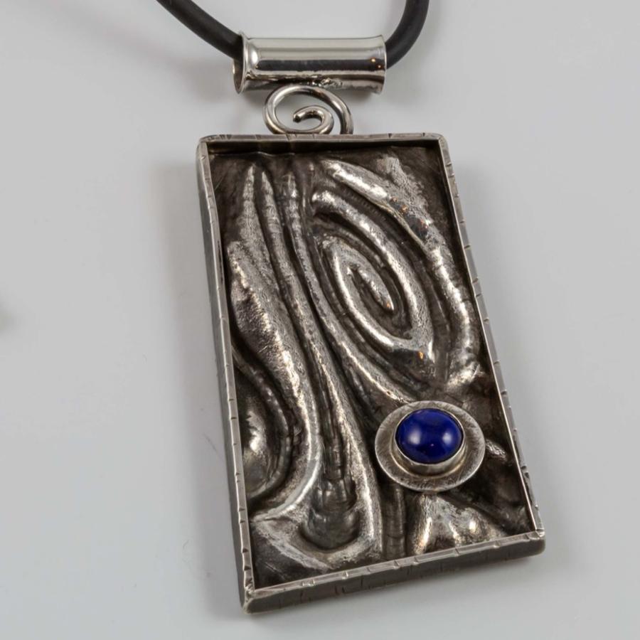 Photograph of a rectangular, textured silver pendant with a small blue stone on a black cord.