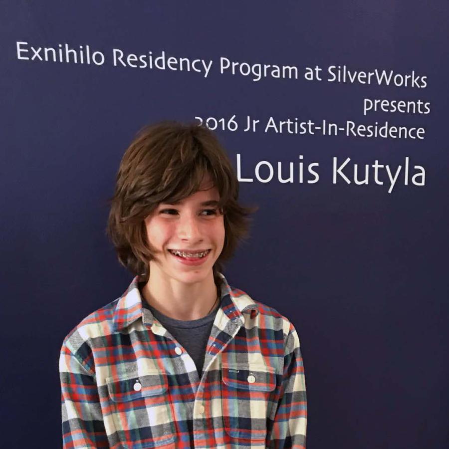 A smiling young person in a plaid shirt stands in front of a blue wall with event signage.