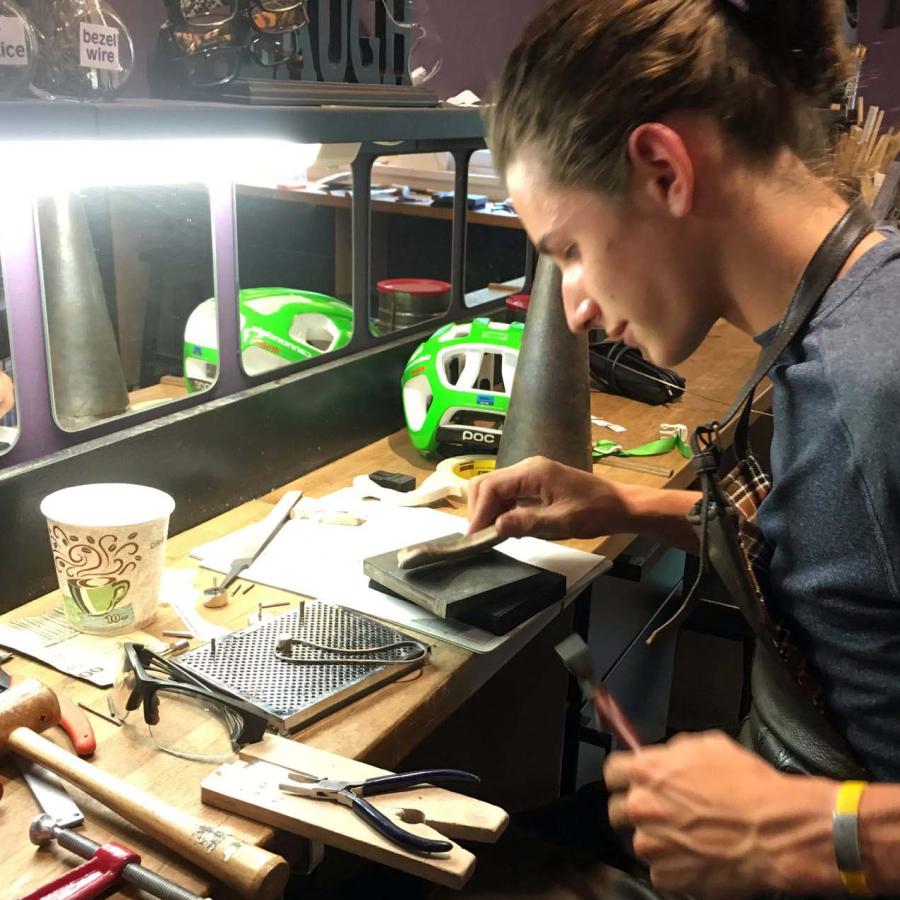 A young artisan focuses on crafting jewelry at a cluttered workbench.