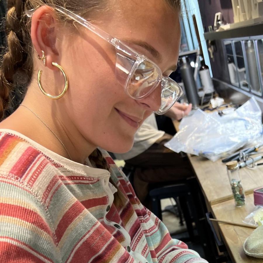 A young woman wearing safety glasses files a piece of metal at a workbench with jewelry tools.