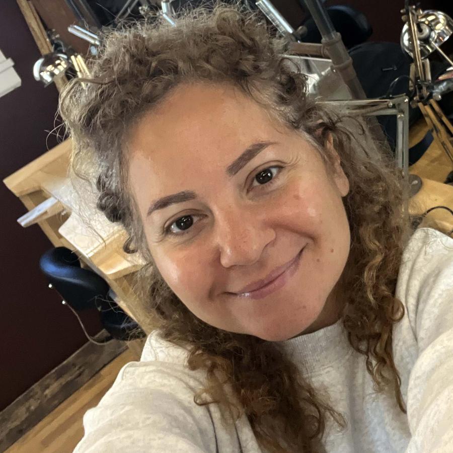 A smiling woman with curly hair takes a selfie in a jewelry workshop.