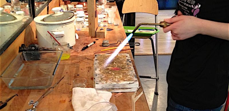 A jeweler works with a soldering torch at a wooden workbench.