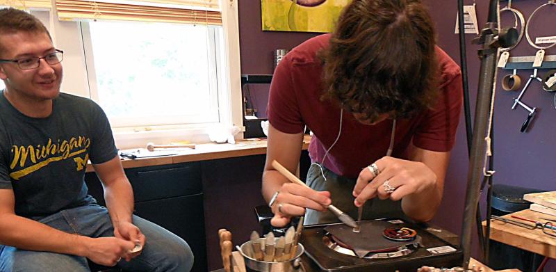 Two people working in a jewelry studio, one focusing on fine detail work at a wooden workbench.