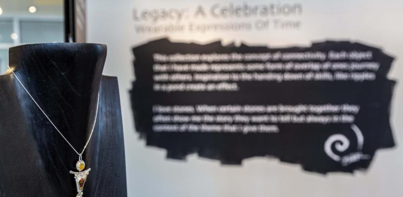 A pendant necklace displayed on a black mannequin in front of a blurry exhibition wall.