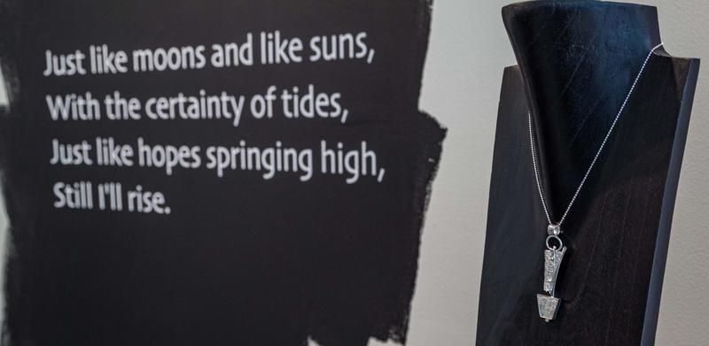 Photograph of a silver pendant necklace on a black stand next to a Maya Angelou quote.