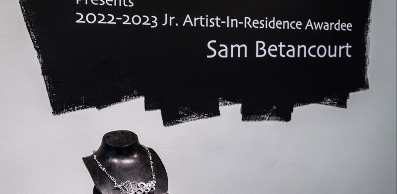 A necklace displayed on a mannequin stand below a sign honoring artist Sam Betancourt.
