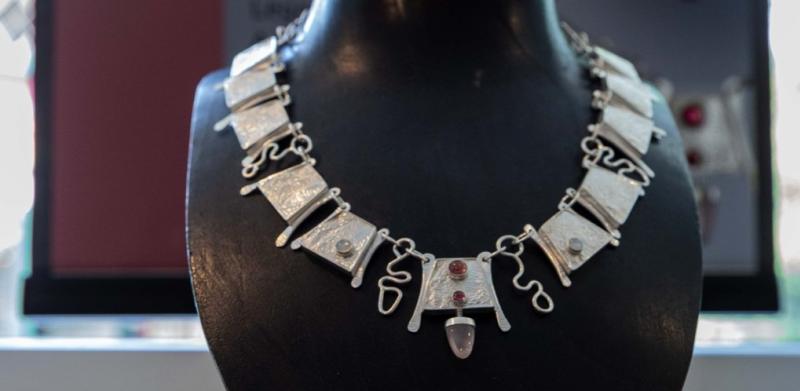 An intricate silver and white inlaid necklace on a display stand in front of an information banner.