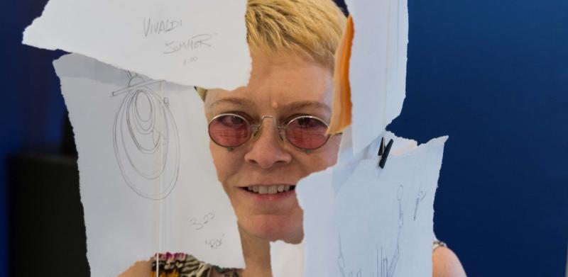 A woman with short blonde hair and glasses poses behind several sketches hanging on strings.