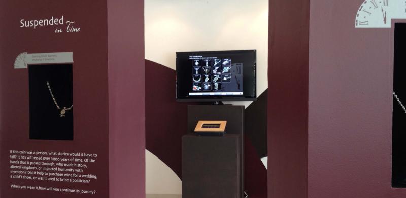 Museum display with jewelry in wall niches and an information screen on a central pedestal.