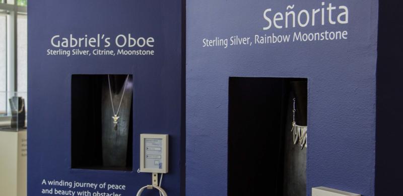 Two navy blue display pillars at a gallery featuring jewelry in recessed niches with headphones.