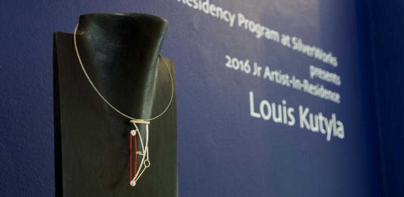 A geometric pendant necklace on a black display stand against a blue gallery wall.