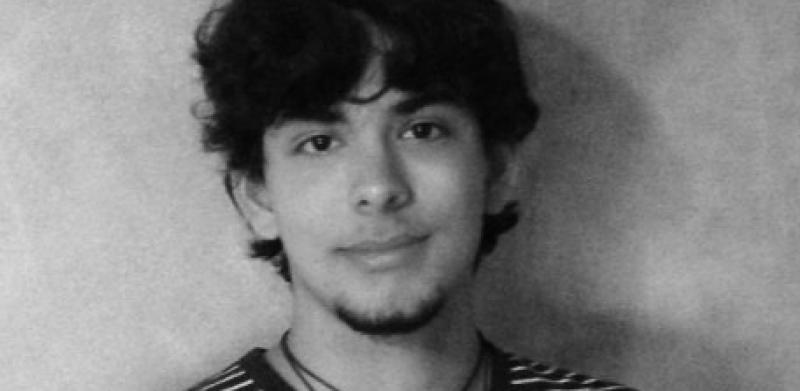 Black and white portrait of a young man with dark curly hair wearing a striped shirt and necklace.