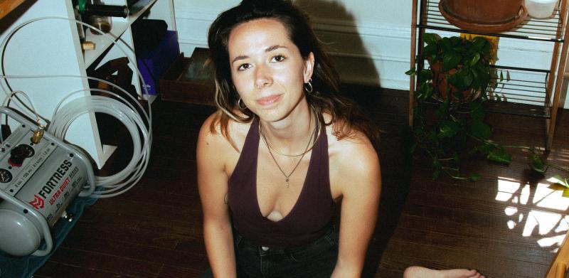 A woman sits on a wooden floor with open notebooks, metal tools, and a pencil around her.