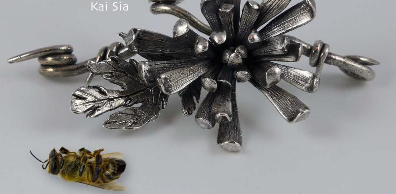 Photograph of a metallic floral brooch next to a dead bee.