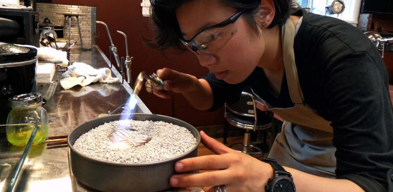 Photograph of a jeweler using a handheld torch to solder metal in a pan of small stones.