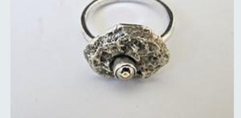 Photograph of a silver ring featuring a textured, organic-shaped setting with a small center stone.