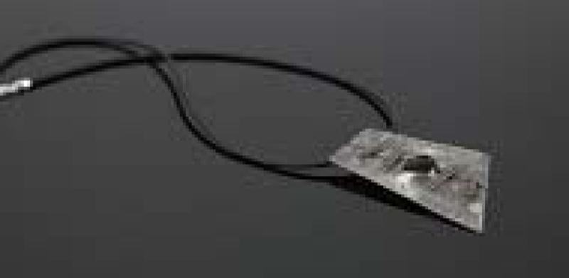 A necklace with a thin, textured, metallic, trapezoid-shaped pendant on a black cord.