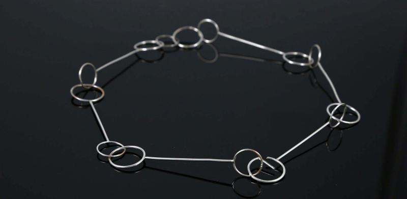 Minimalist silver necklace made of linked thin metal rods and rings on a dark background.