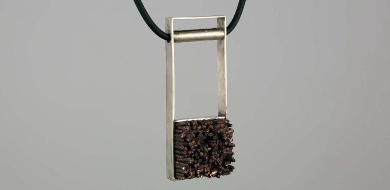 Photograph of a silver, rectangular pendant with a textured dark brown base on a black cord.