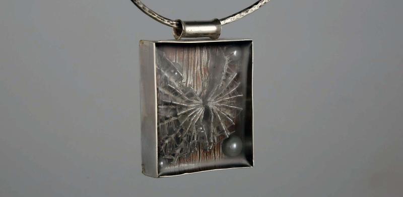 Photograph of a rectangular silver pendant containing a textured gray element and a small dark bead.