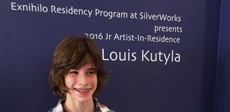A smiling young person in a plaid shirt stands in front of a blue wall with event signage.
