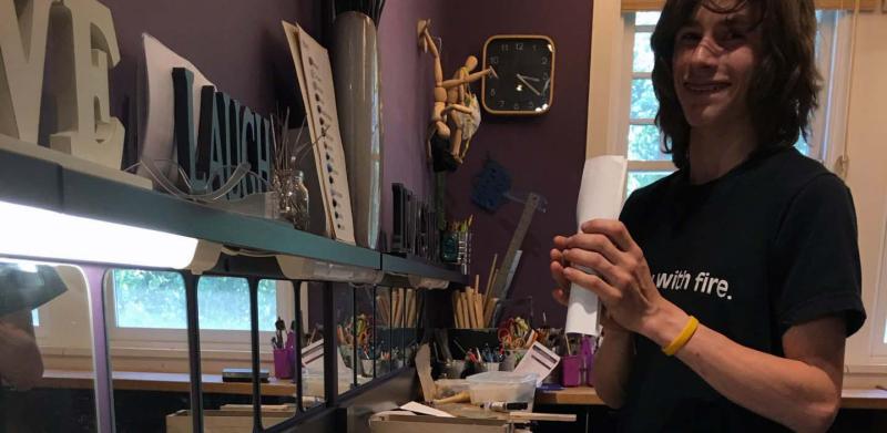 A person stands behind a workspace counter lined with small crafts and paper tags in a studio.