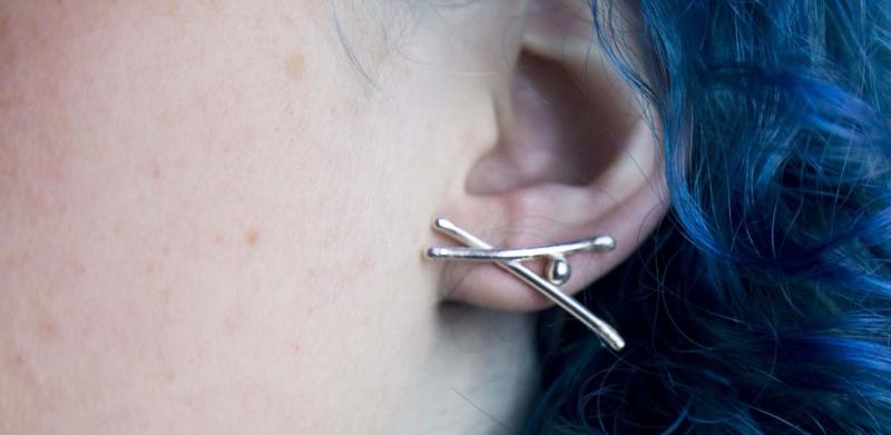 Close-up photograph of a person with blue hair wearing a silver, X-shaped stud earring.