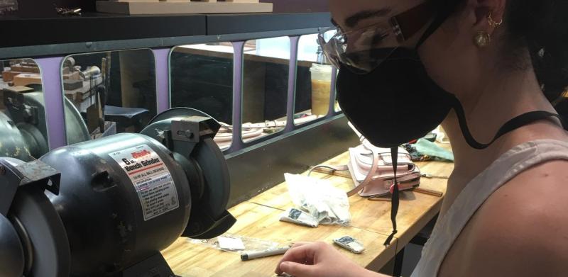 A masked woman grinds pieces of metal at a workbench.