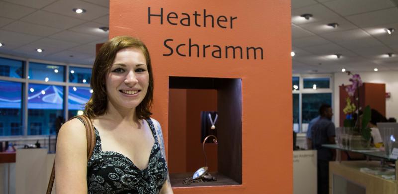 Photograph of a woman smiling, standing beside an orange pillar with the name Heather Schramm.
