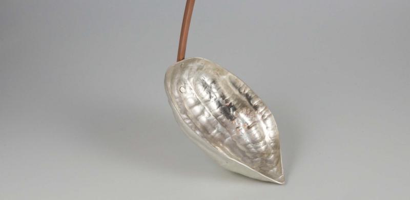 Photograph of a silver ladle with a copper handle, on a neutral gray background.