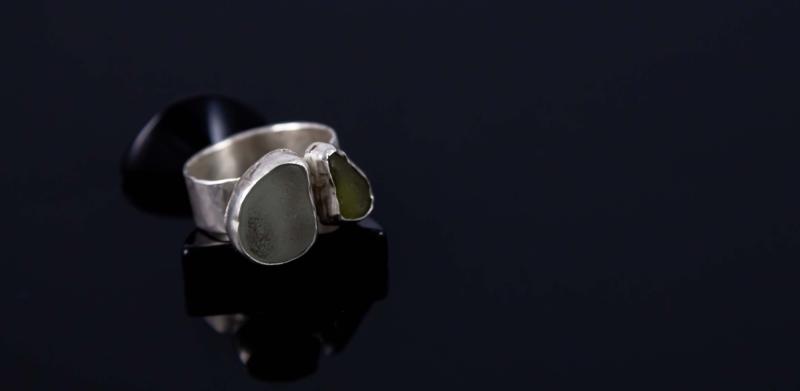 A silver ring with two sea glass stones on a dark reflective surface.
