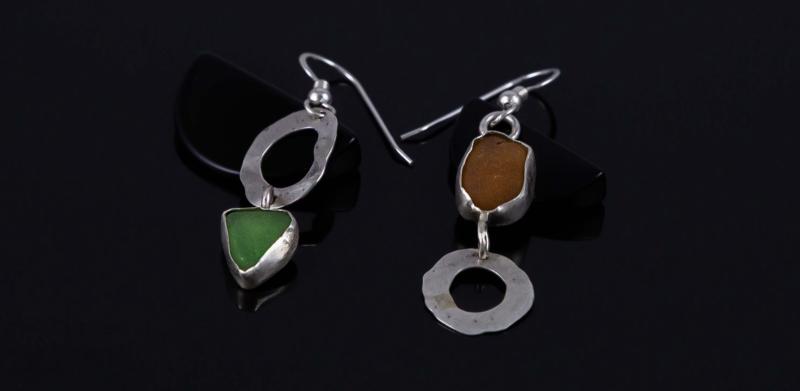 A photograph of two asymmetrical earrings with sea glass and silver on a dark background.