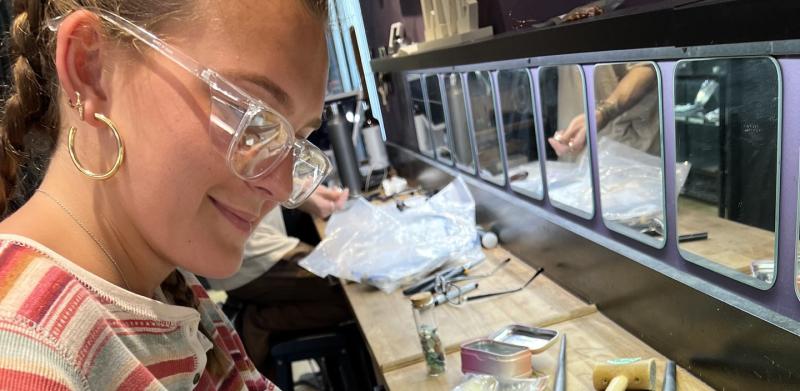 A young woman wearing safety glasses files a piece of metal at a workbench with jewelry tools.