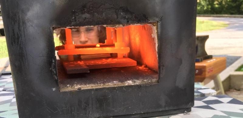 A person's face peers through the glowing, fiery opening of a black metal forge.